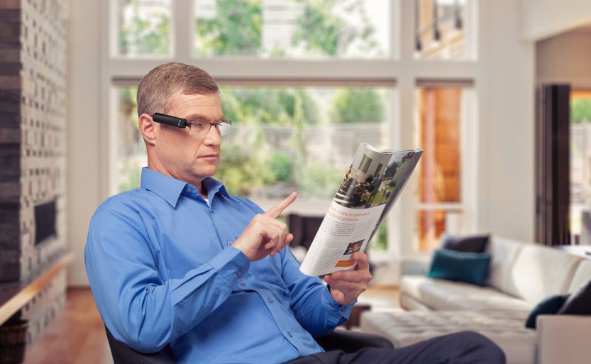 Das Bild zeigt die OrCam Brillenkamera im Einsatz. Ein Mann lässt sich per Fingerzeig ein Magazin vorlesen.